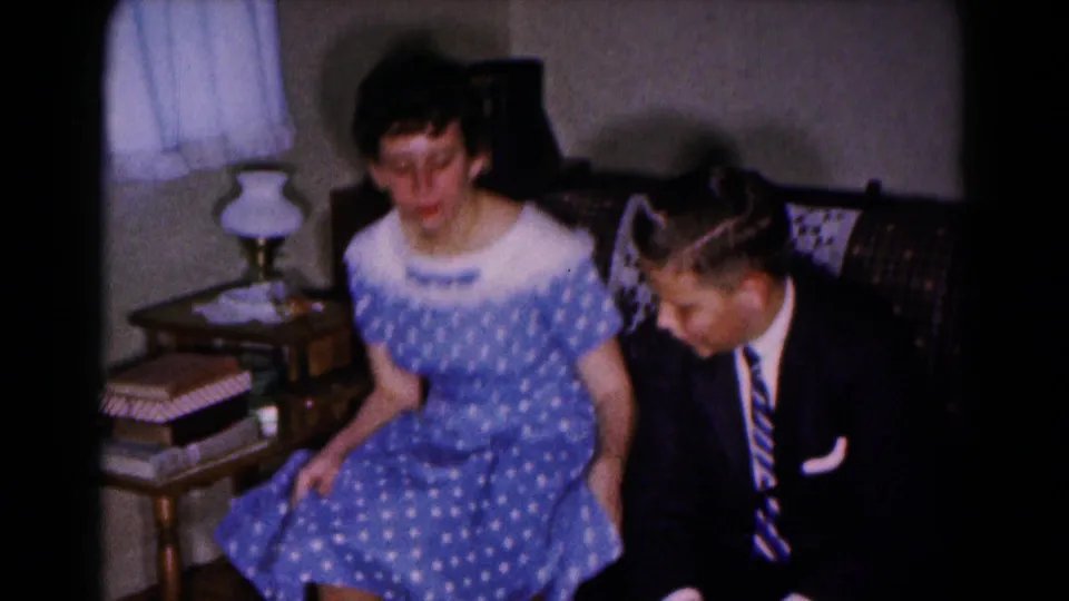 The woman sits beside the suited man on a patterned couch in a 1961 Hagerstown home interior as both look downward.