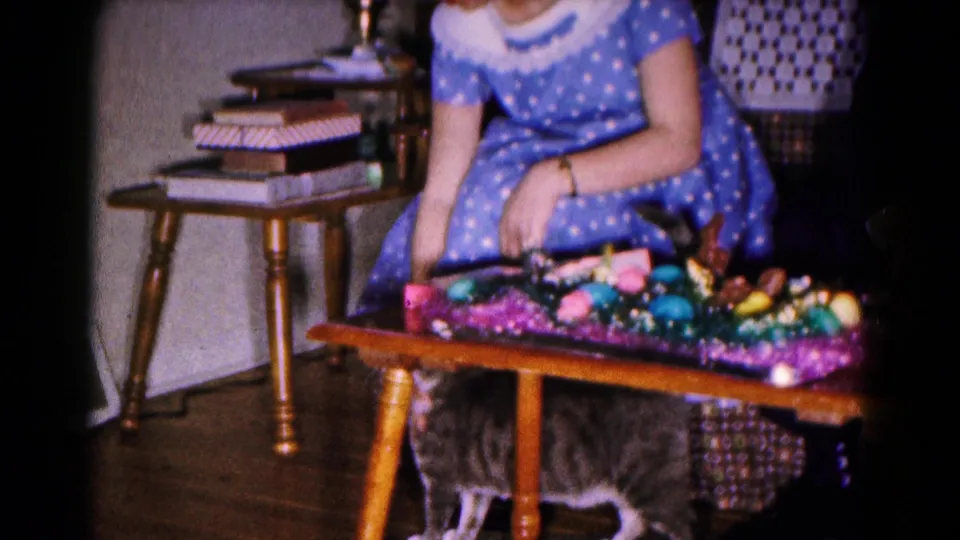 1961 Hagerstown home interior where a woman in a blue polka dot dress reaches down toward a cat beside an Easter table.