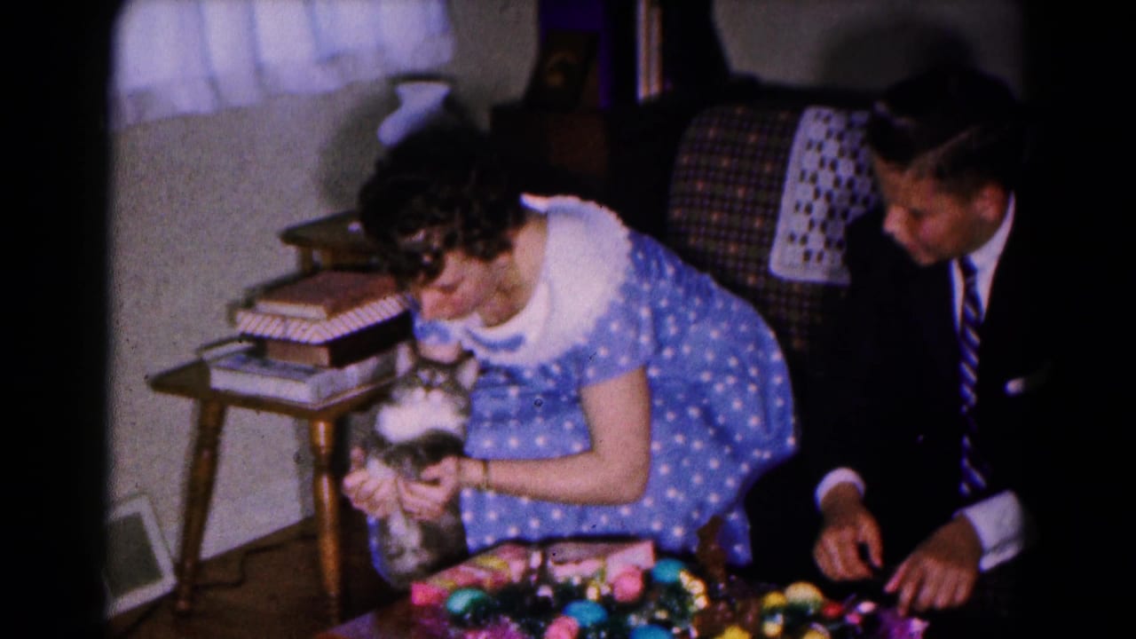 Woman in a blue polka-dot dress holds a small cat beside a suited man and Easter decorations.