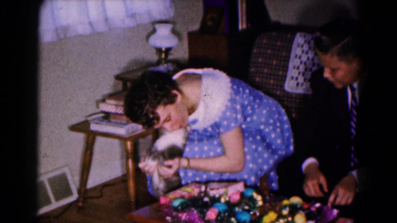 1961 Maryland home-movie frame of a woman leaning toward a cat beside Easter decorations, with a suited man seated in the living room.