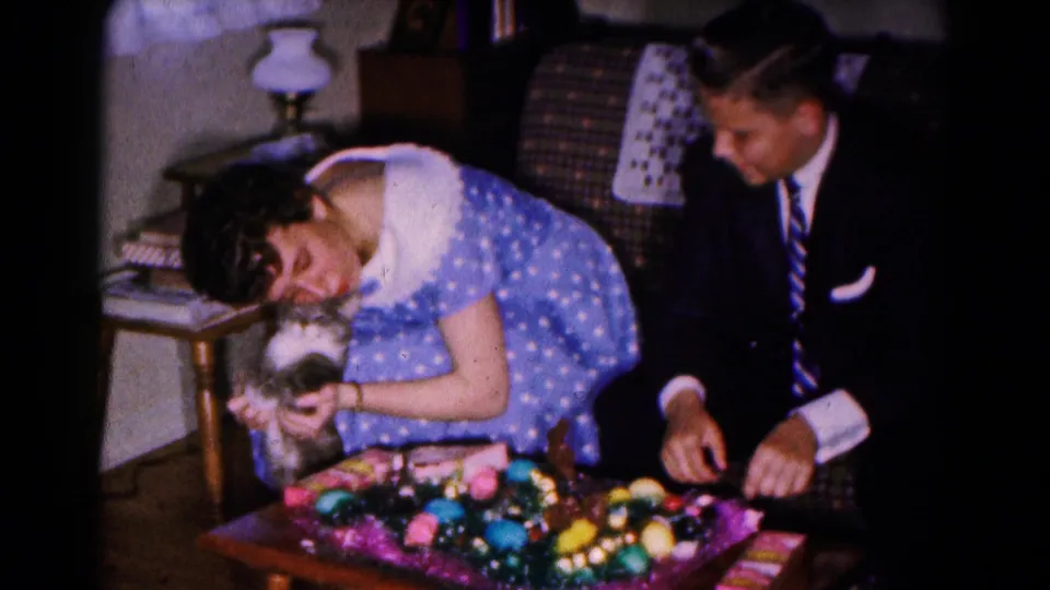 Indoor 1961 Easter home-movie frame with a woman cradling a cat, a suited man beside her, and colorful decorations in the foreground.