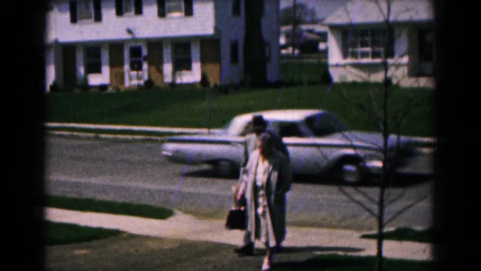 1961 Hagerstown frame showing two adults arriving along a residential sidewalk, with houses and a parked car in the background.