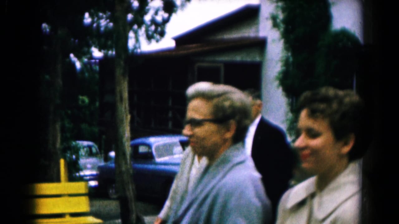 1961 Hagerstown frame with adults near trees, a yellow bench or barrier, and parked mid-century cars outside a home.