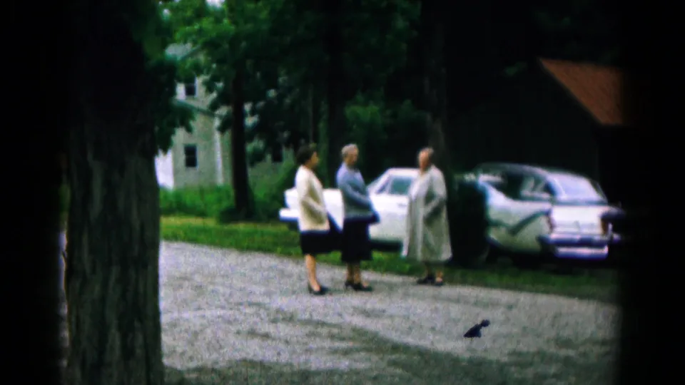 1961 Hagerstown home-movie frame showing three women conversing in a drive beside light-colored mid-century cars.