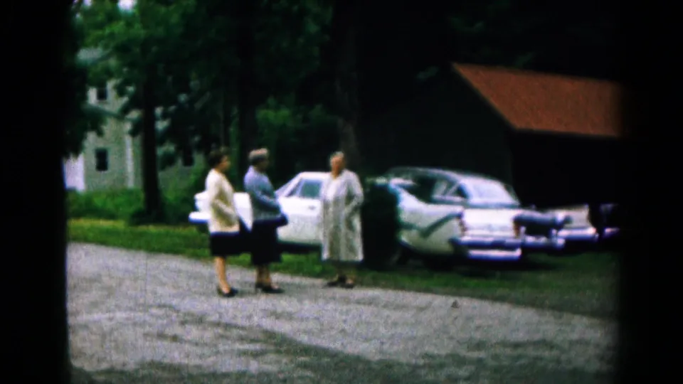 1961 Hagerstown frame of three women in a residential drive near parked mid-century cars and shaded trees.