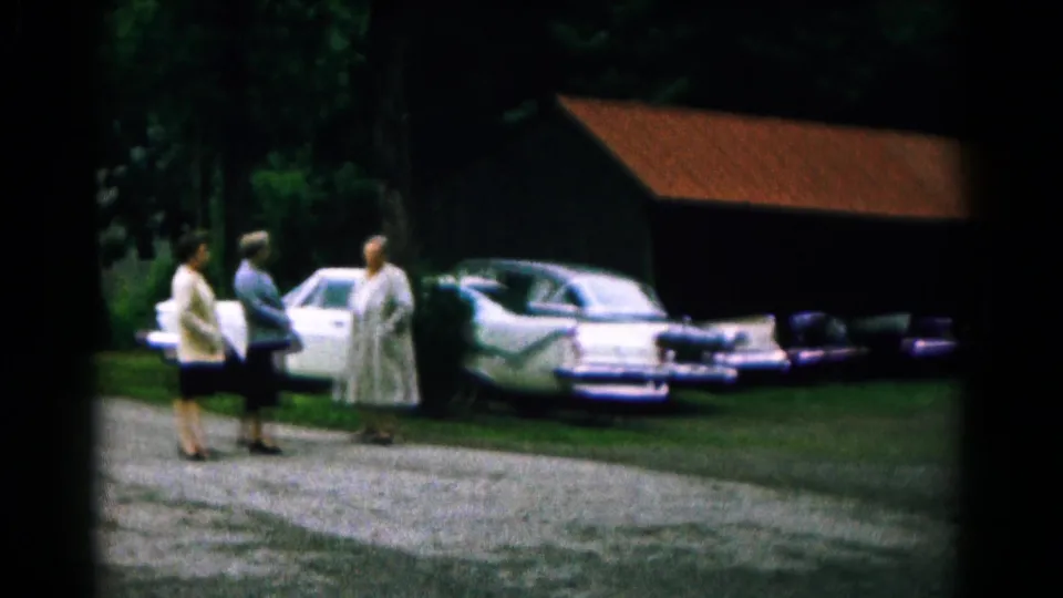In 1961 Hagerstown, Maryland, three women talk beside mid-century parked cars near trees and a red-roofed barnlike structure.