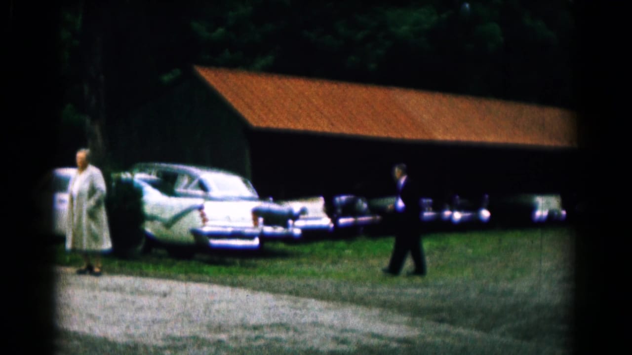 A man in a dark suit walks toward women standing by parked cars and a red-roofed structure.