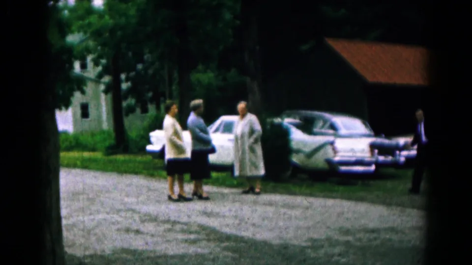 The closing frame shows the women gathered on the gravel drive with parked cars, trees, and the red-roofed structure behind them.