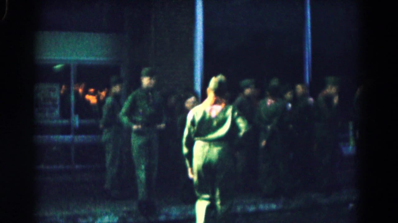 1961 Hagerstown frame with a soldier in green uniform and red neckerchief walking past other soldiers near a lit entrance.