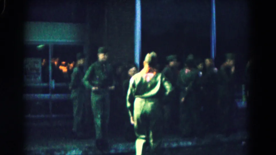 1961 Hagerstown frame with a soldier in green uniform and red neckerchief walking past other soldiers near a lit entrance.