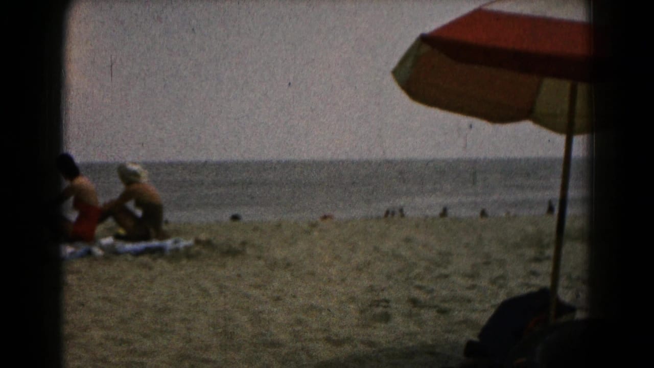 Two women sit on towels on a sandy beach near the water, with a beach umbrella at the right edge.
