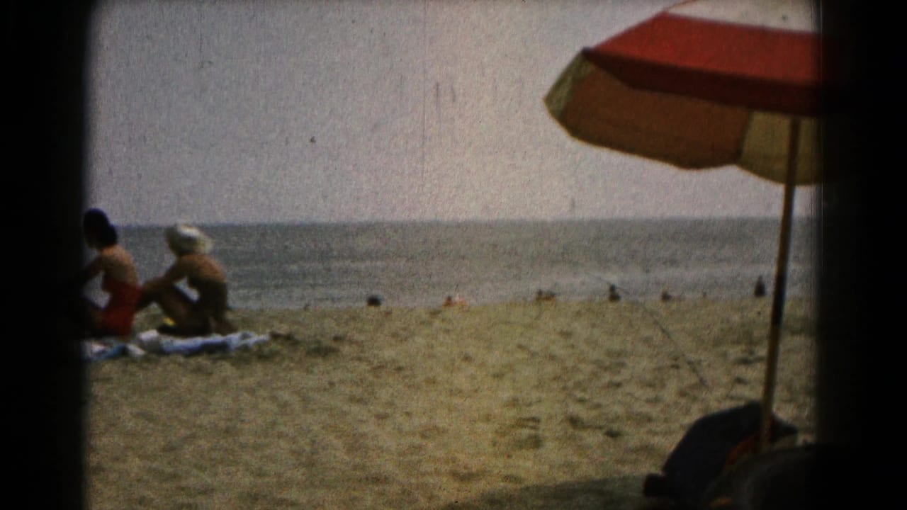 Two women remain seated on towels by the shore, with distant bathers and an umbrella visible.