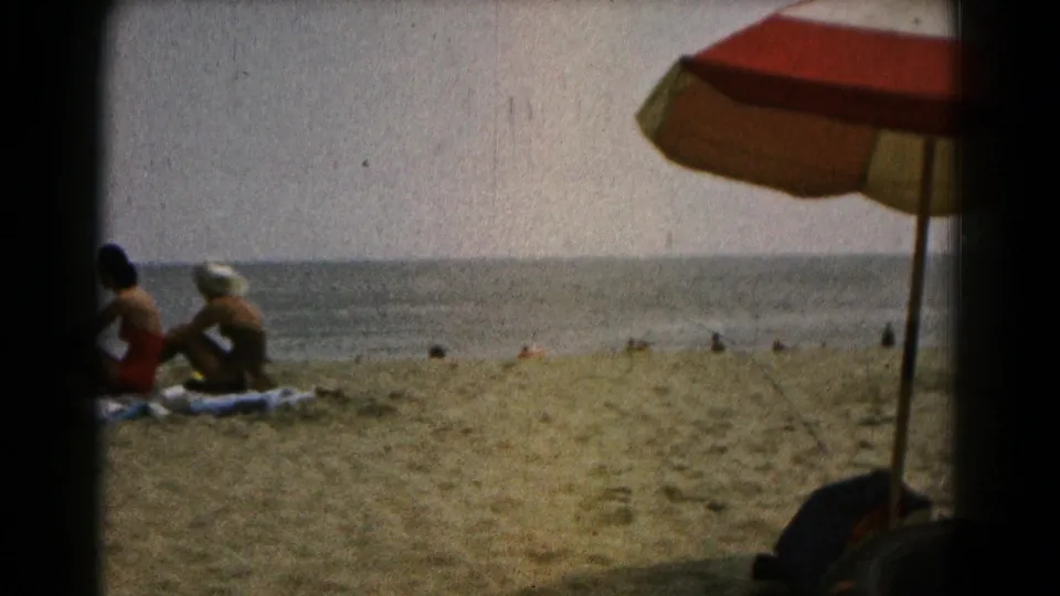 1961 beach home-movie still showing two women seated on towels near the water, with distant beachgoers and a striped umbrella.