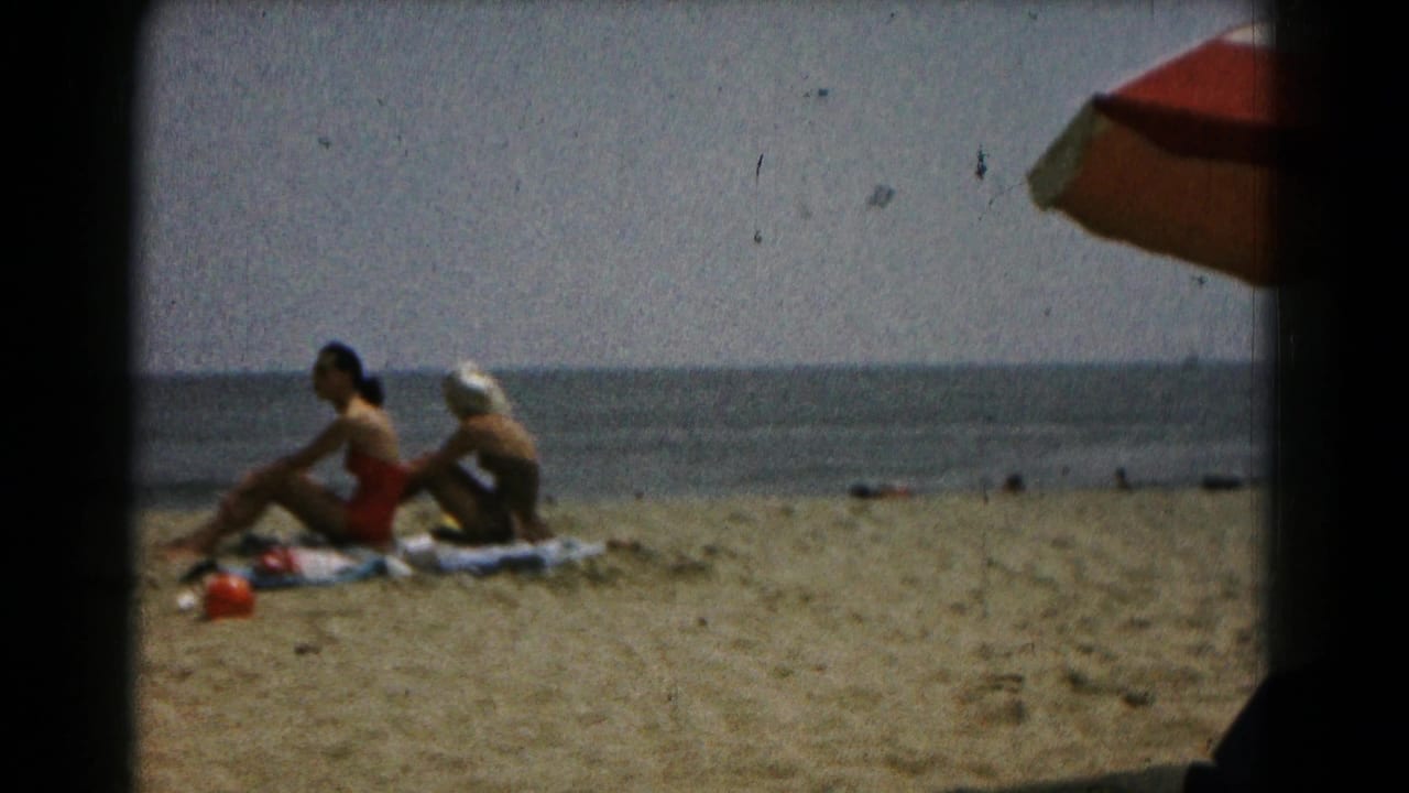 1961 8mm frame of two adult women seated on towels on a sandy beach, with water beyond and umbrella fabric at the top right.