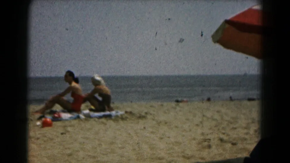 1961 8mm frame of two adult women seated on towels on a sandy beach, with water beyond and umbrella fabric at the top right.