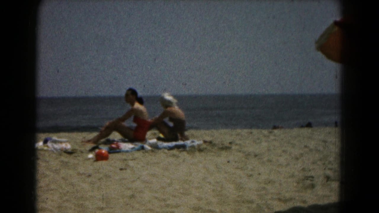 1961 home-movie still of two women relaxing on towels at the beach, with a dark frame edge and calm water in the background.