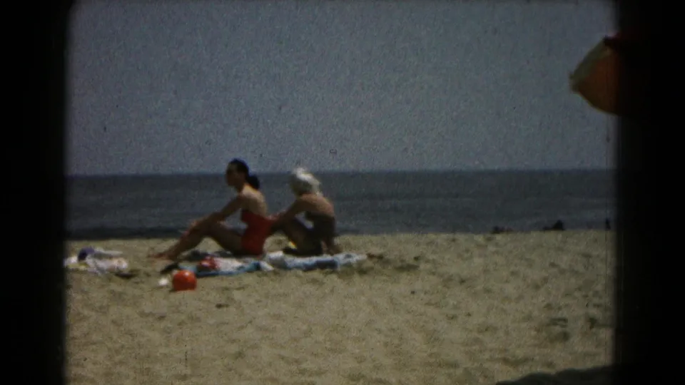 1961 home-movie still of two women relaxing on towels at the beach, with a dark frame edge and calm water in the background.