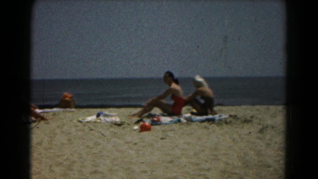 Women remain seated on towels with the ocean behind them and beach items nearby.