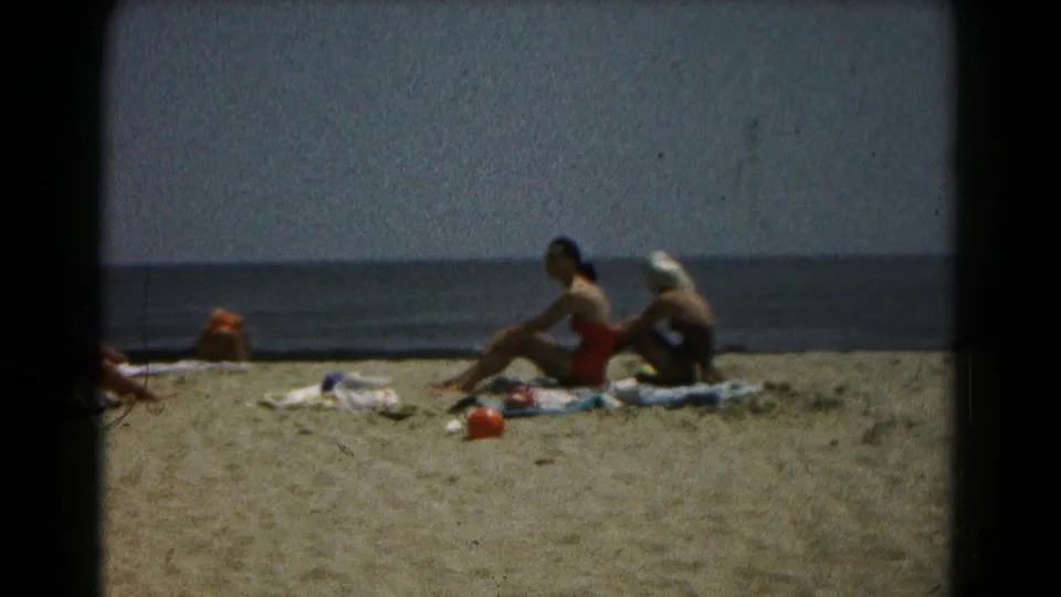 Women in swimsuits sit on towels near the shoreline in supplied 1961 Maryland home-movie footage.