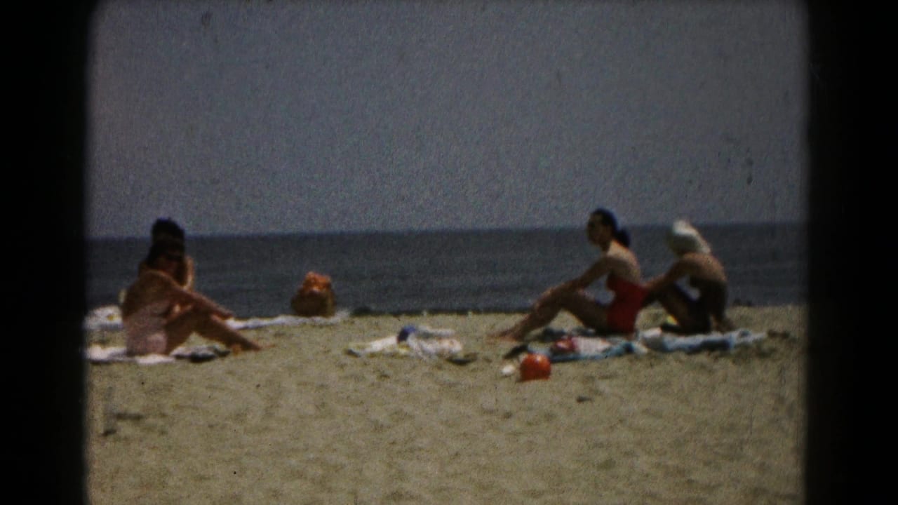 Home-movie beach frame with a foreground woman seated on a towel and two women farther right near the ocean.