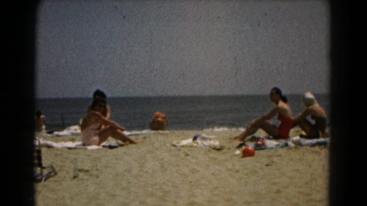 1961-style 8mm frame showing women sunning and talking on a sandy beach with the ocean in the background.