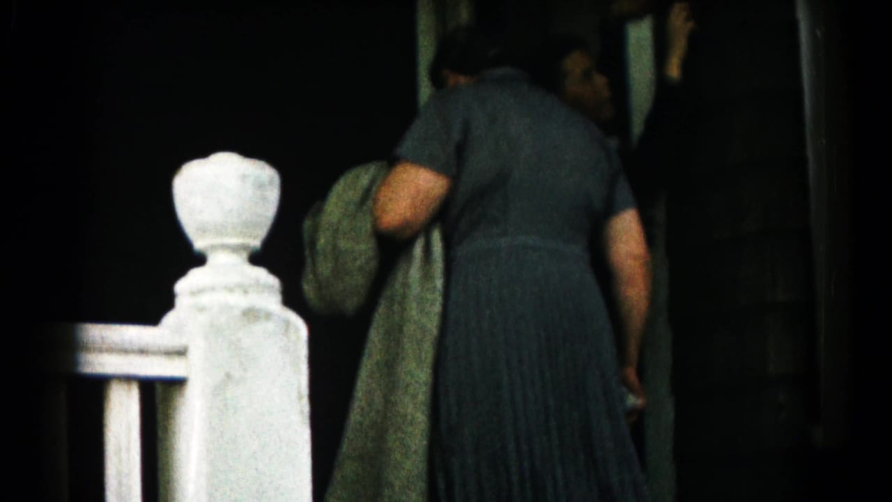A woman in a dark dress stands at a doorway with a coat over her arm.