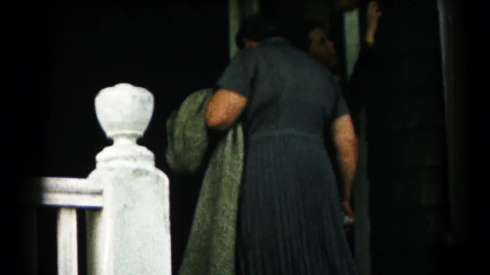 A woman in a dark pleated dress stands near a house entrance, with another adult partly visible inside the doorway.
