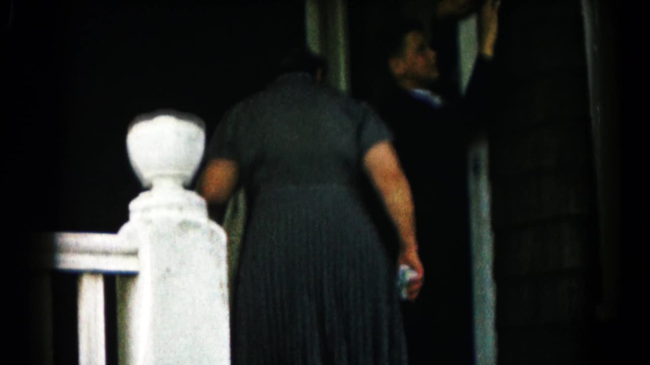 The 1961 home-movie frame shows a woman entering a house, seen from behind near a white porch post and dark doorway.