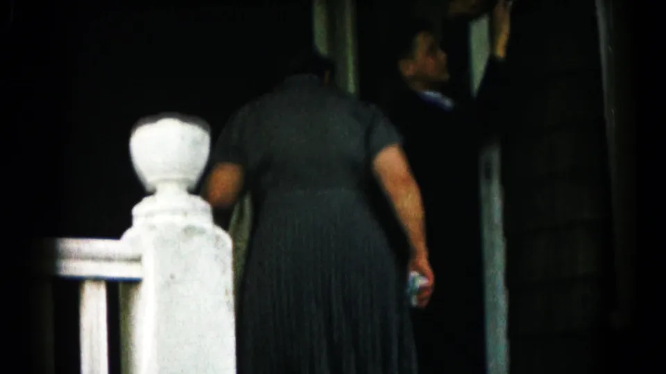 The 1961 home-movie frame shows a woman entering a house, seen from behind near a white porch post and dark doorway.
