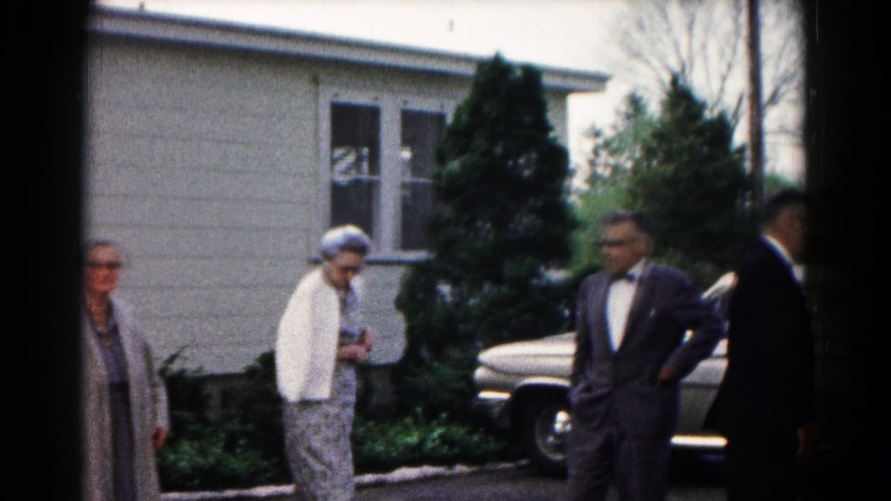 In 1961 Hagerstown, Maryland home-movie footage, adults in suits and coats stand outside a suburban house near a parked sedan.