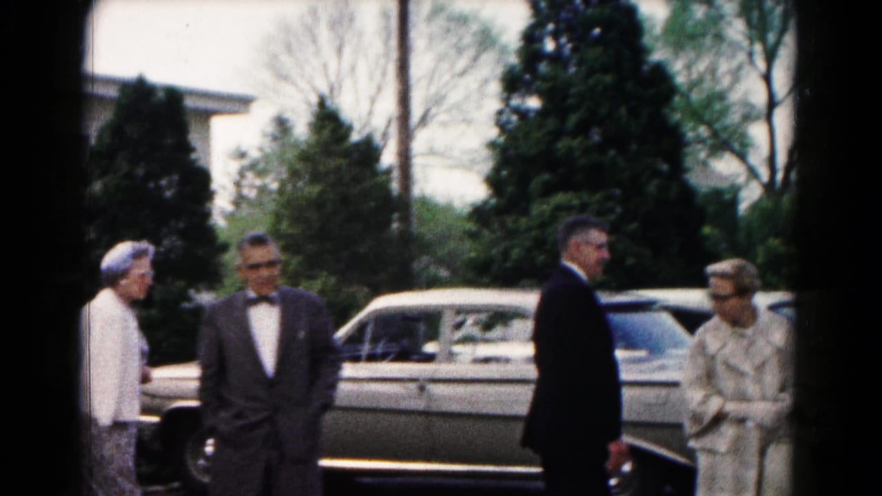 Well-dressed adults stand near a light-colored sedan on a residential street.