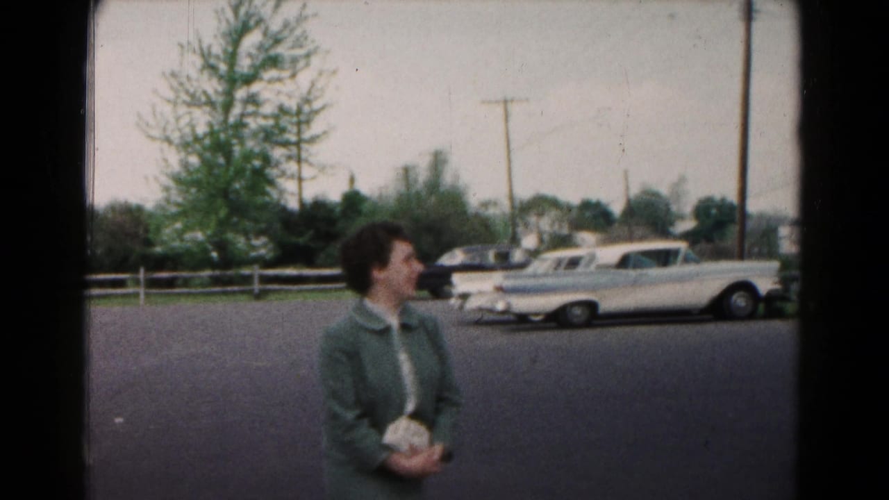 A woman in a green coat stands near a residential street with parked mid-century cars in the background.
