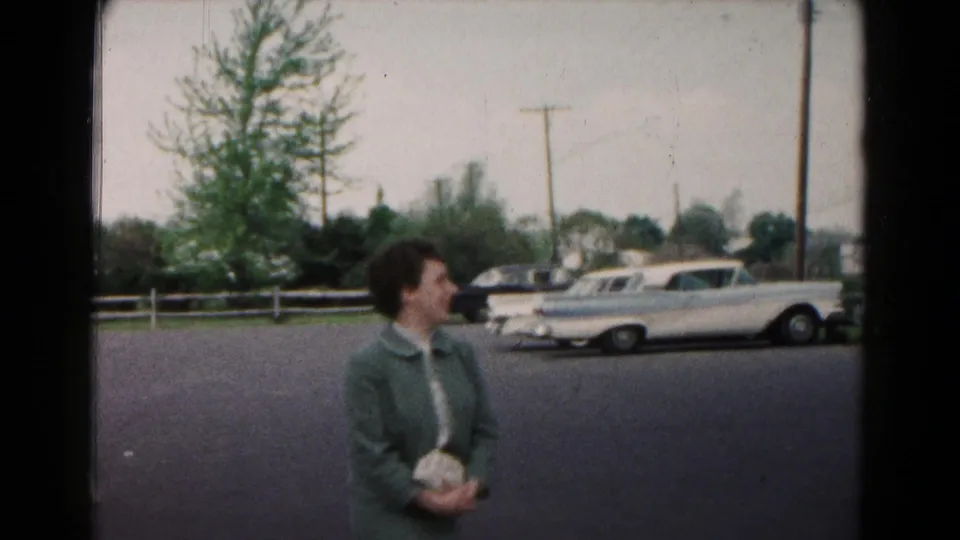 A woman in a green coat stands near a residential street with parked mid-century cars in the background.