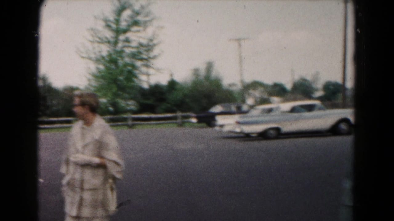 A woman in a light coat appears near a residential street with parked cars in 1961 Hagerstown, Maryland home-movie footage.