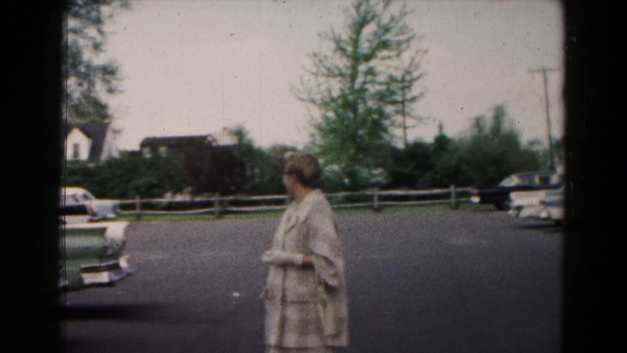 In 1961 Hagerstown home-movie footage, a woman in a light coat stands in a paved lot near mid-century cars, trees, and houses.