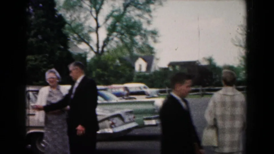 Adults in formal wedding attire stand and walk near parked cars in the 1961 Hagerstown parking-lot scene.