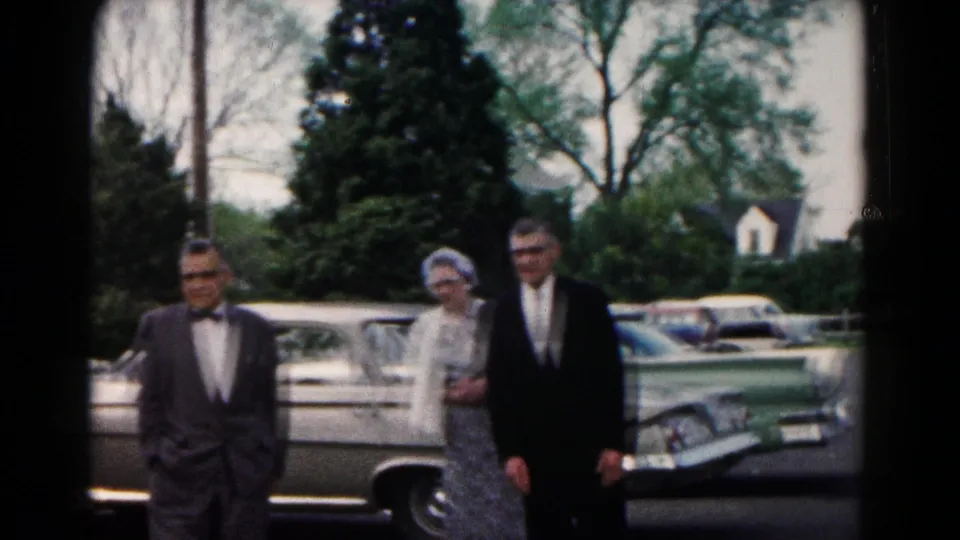 Two men in suits and a woman in a dress appear near a parked car in the 1961 Hagerstown wedding gathering footage.