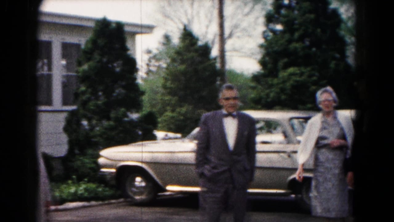 A suited man walks past a parked mid-century car as a formally dressed woman stands nearby in the 1961 Hagerstown scene.