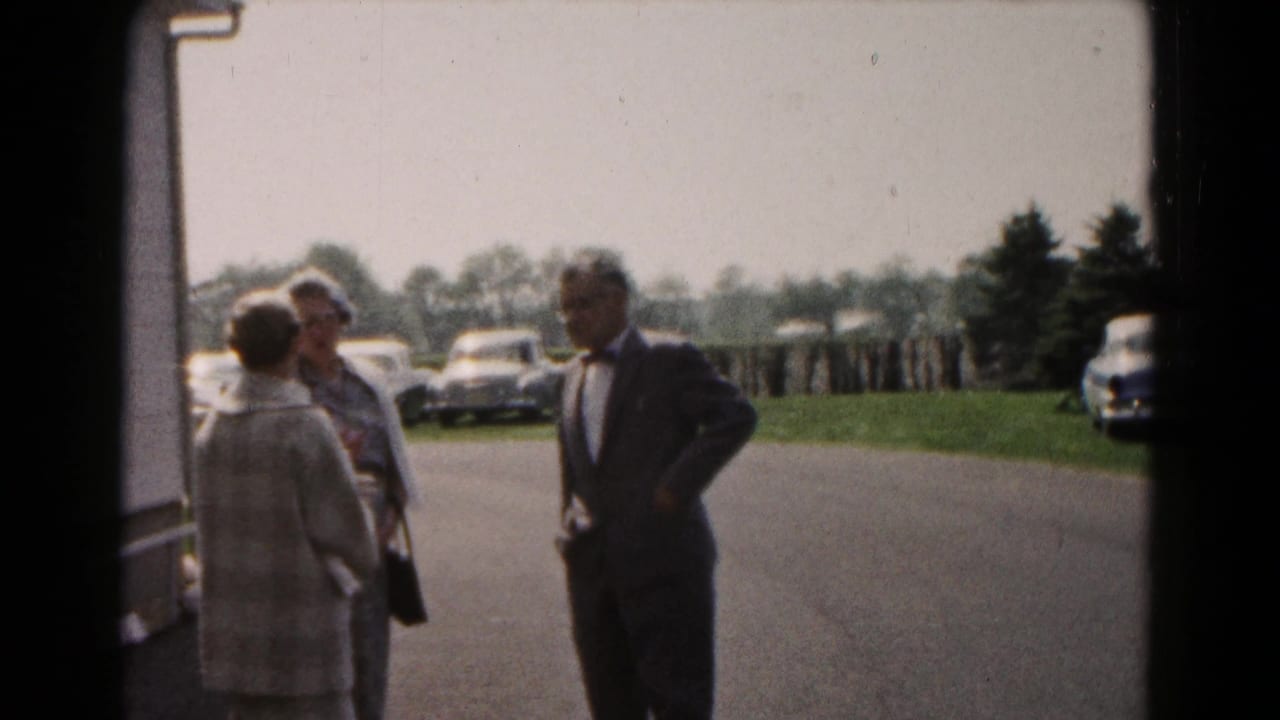 In 1961 Hagerstown home-movie footage, two women and a suited man stand by a building near a parking lot with cars in the background.