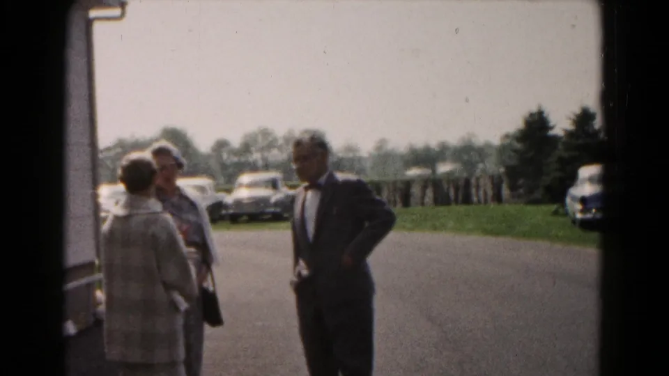 In 1961 Hagerstown home-movie footage, two women and a suited man stand by a building near a parking lot with cars in the background.