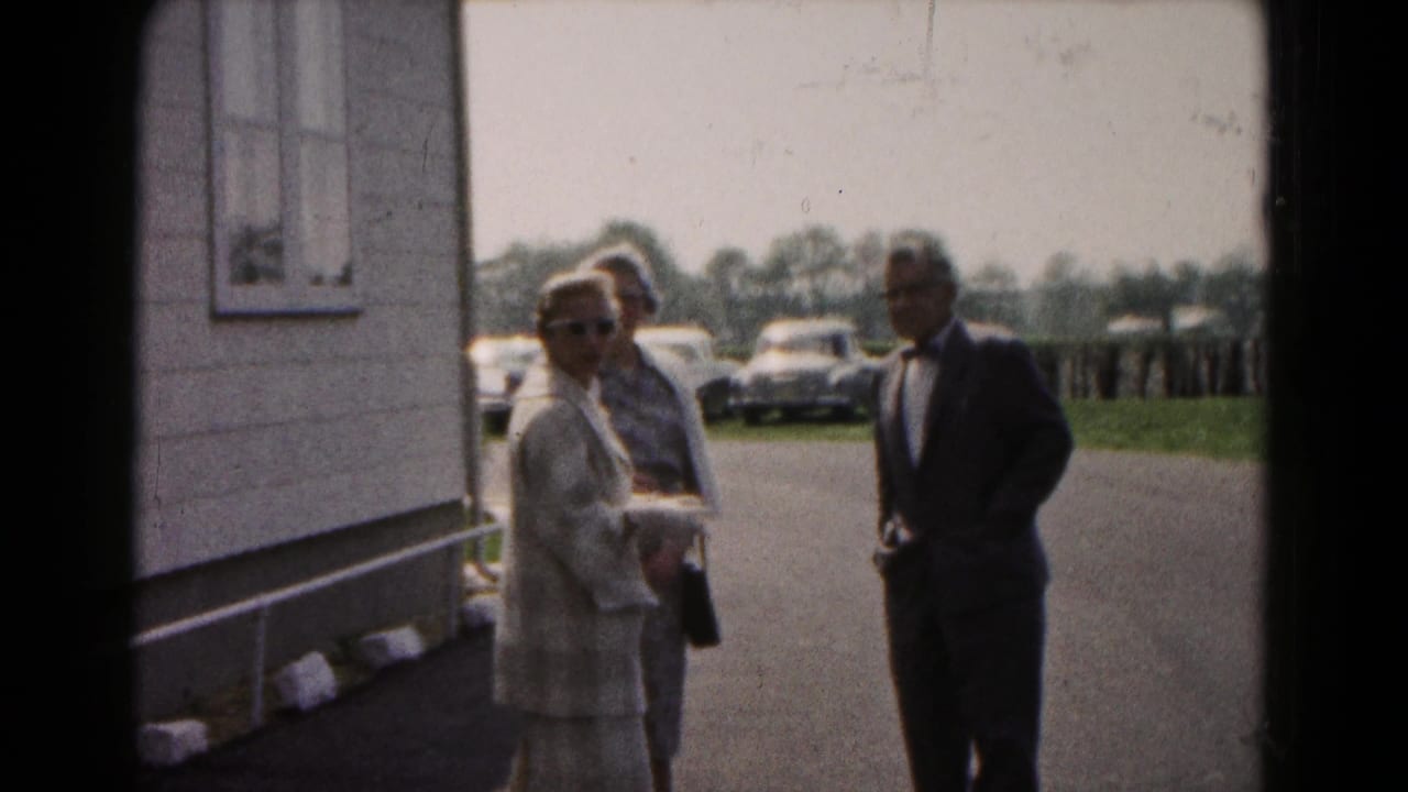 Adults stand beside a building while several cars are parked in the background.