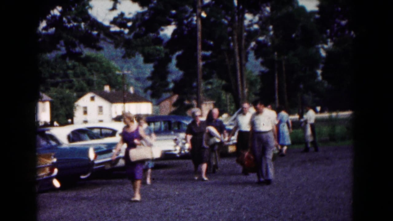 Several adults walk through a Hagerstown parking lot carrying luggage, with classic cars, trees, and houses in the background.