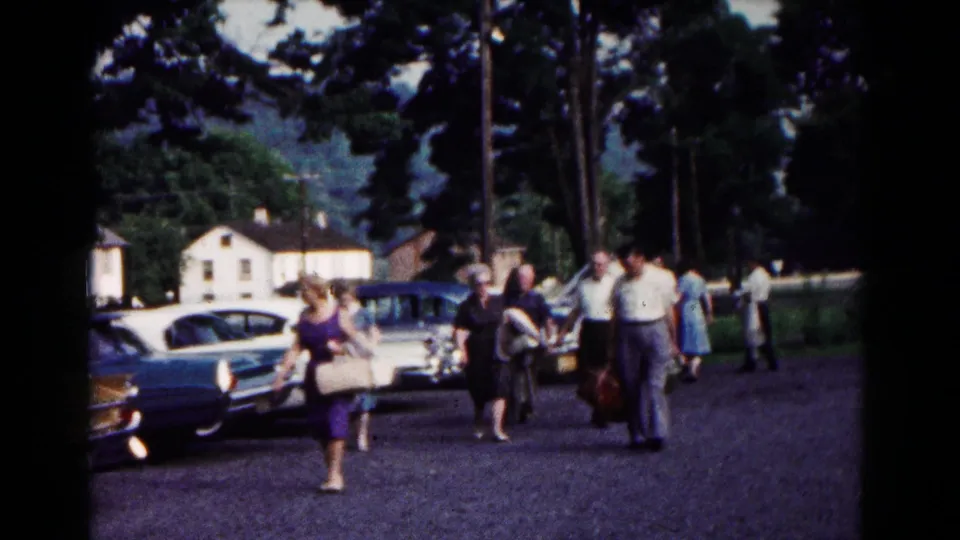 Several adults walk through a Hagerstown parking lot carrying luggage, with classic cars, trees, and houses in the background.