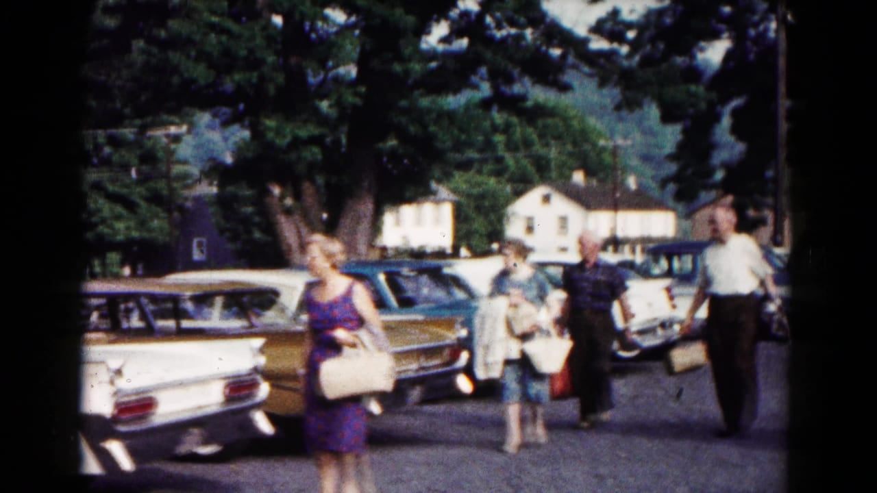 Adults carrying bags pass parked sedans in 1961 Hagerstown footage, with trees and nearby houses visible beyond the lot.