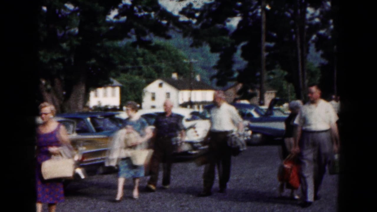 Adults walking through a parking area with classic cars and trees behind them.