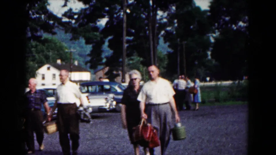 Park visitors in 1960s attire carry bags and baskets through a Hagerstown parking area with cars and trees in the background.