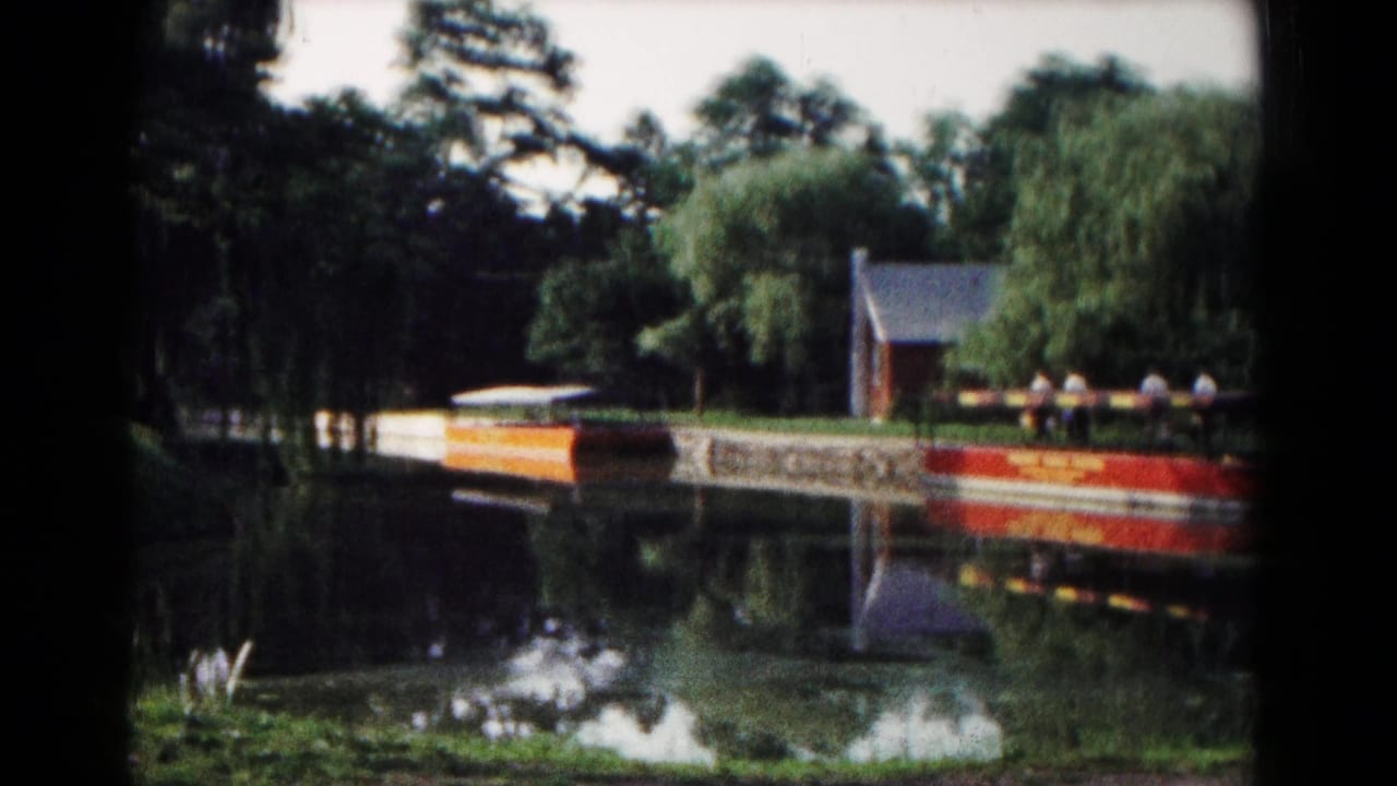 The Hagerstown park lake reflects trees, a small lakeside building, and a red-and-white dock or boat in soft 8mm color.
