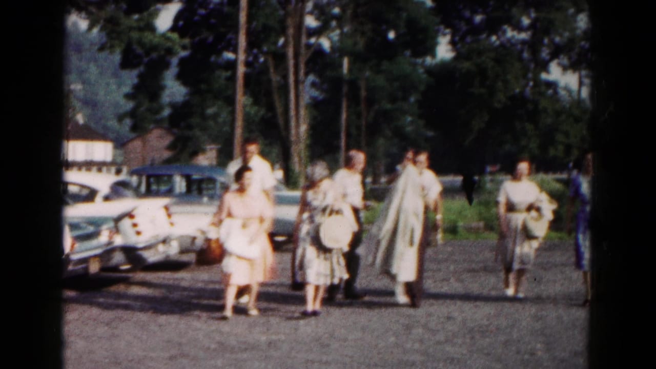 Adults walk through a gravel parking area near parked mid-century cars and trees.
