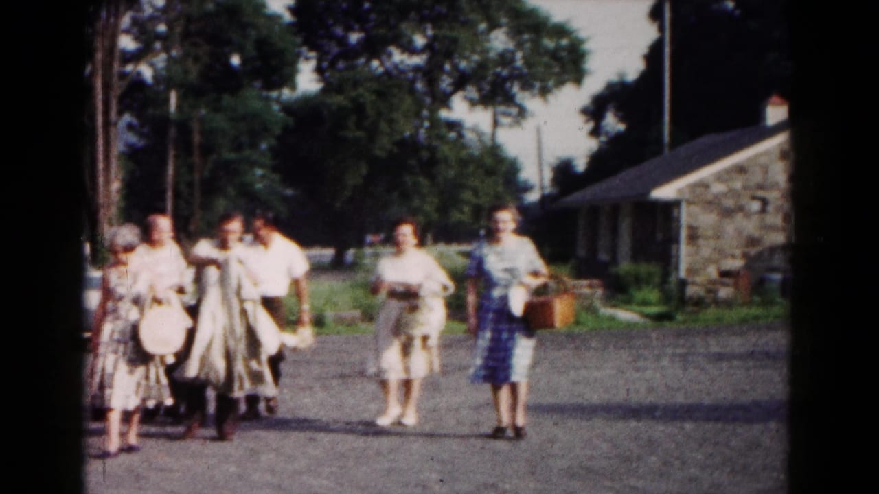 Adults in mid-century clothing approach the camera in a gravel area near a stone building and trees.