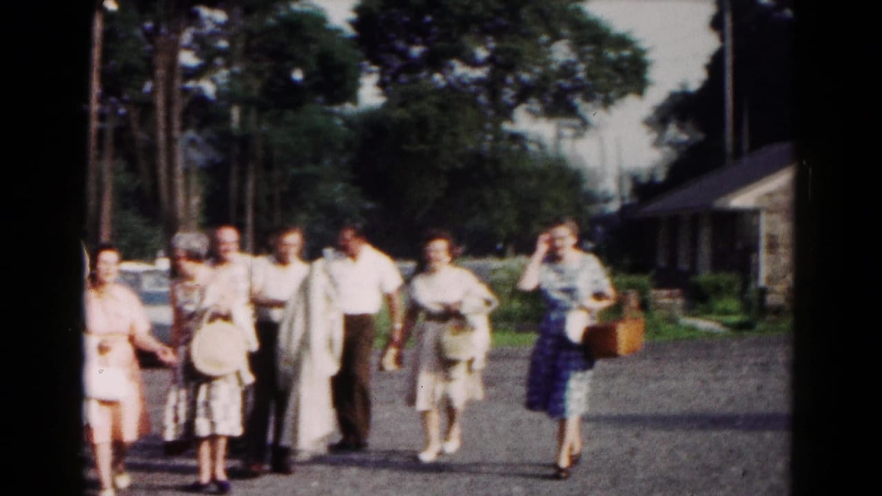 A mixed group of adults walks toward the camera in a 1961 Hagerstown lakeside parking area with trees and cars behind them.